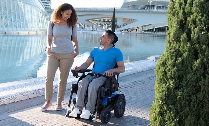 Eine junge Frau und ein Mann im Rollstuhl gehen in städtischer Umgebung spazieren. Der Mann nutzt einen E-Rollstuhl. Copyright: Invacare