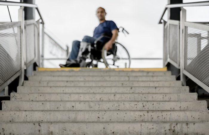 Ein Mann im Rollstuhl steht vor einer Treppe, die nach unten geht. Der Mann ist nur verschwommen zu sehen. 