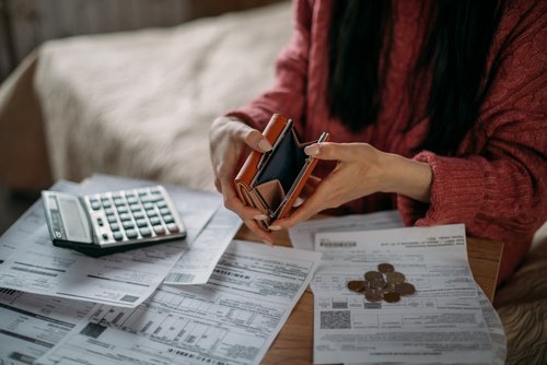 Eine Frau in einem roten Pulli sitzt vor Rechnungen und einem Taschenrechner. Sie hält ihre leere Geldtasche offen und man sieht, dass kein Geld darin ist. 