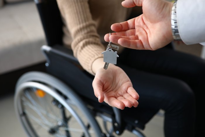 Das Bild zeigt eine Person im Rollstuhl, der ein Schlüssel übergeben wird. An dem Schlüssel hängt ein Anhänger in Form eines Hauses. 